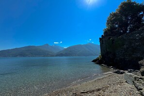 Vista despejada de un lago con montañas al fondo, una orilla rocosa y un muro de piedra a la derecha.