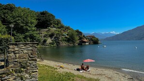 A lakeside scene with a stone wall, a person under an umbrella, and boats on the water. - Villetta Alessia in Menaggio (San Siro)