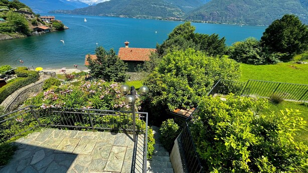 A scenic view of a lake with mountains in the background, a small building with a red roof, and a well-maintained garden area with a stone staircase. - Villetta Alessia in Menaggio (San Siro)
