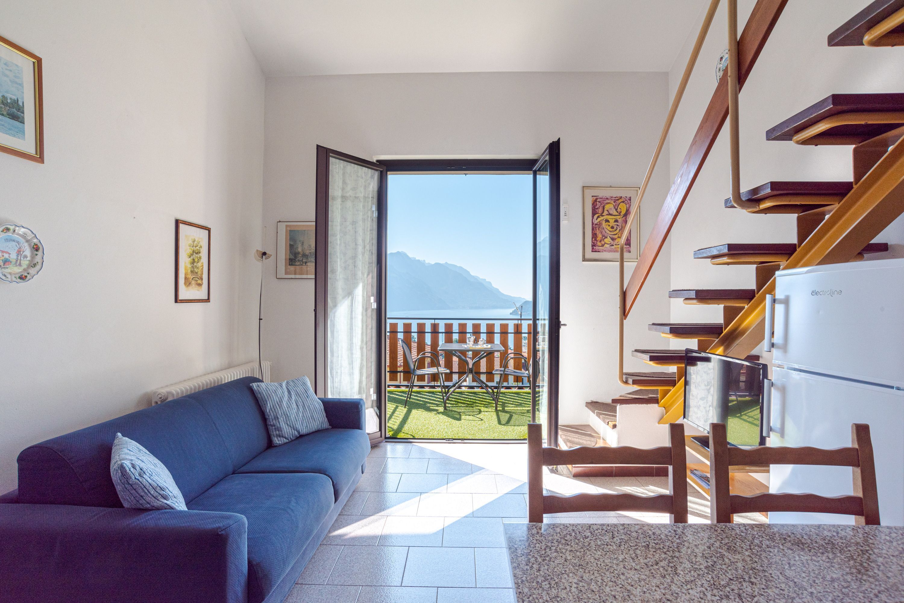 A living room with a blue sofa, a wooden staircase, and a view of mountains through an open door.