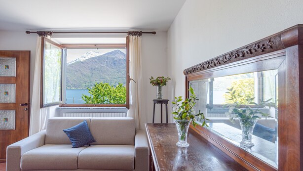 Ein Zimmer mit einer Couch, einem Holztisch mit einer Vase, einem Fenster und Blick auf die Berge sowie einem Spiegel.