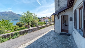 Ein gepflasterter Weg führt zu einem Gebäude mit Tür und Fensterläden, mit Blick auf ein Gewässer und Berge.