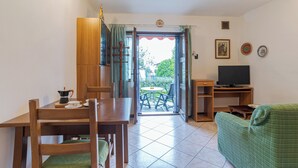 A room with a wooden dining table, chairs, a green sofa, a television, and a view of a garden through an open door. - CO RI Appartamenti Dependance (Menaggio)