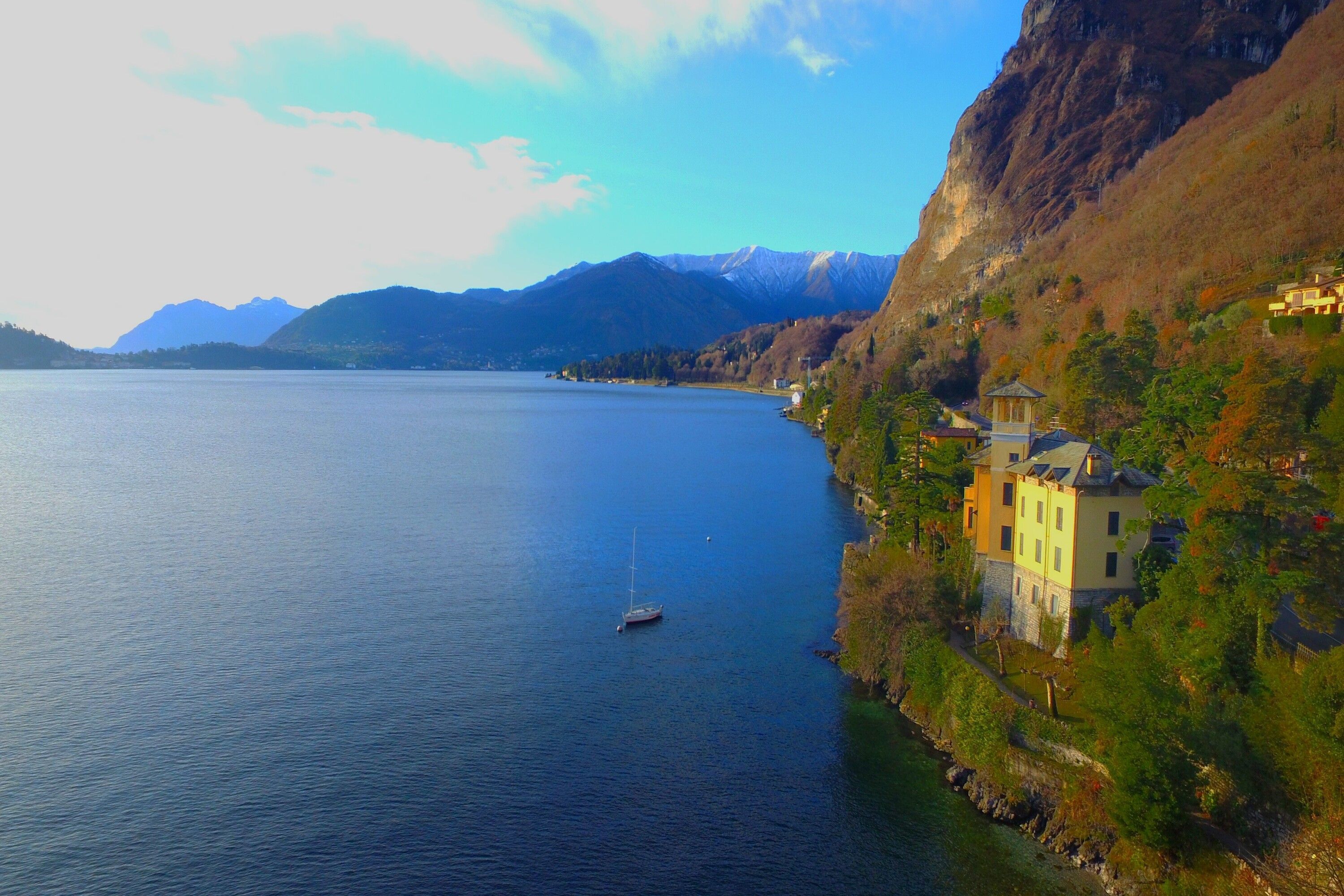 Vista sul lago con una piccola barca a vela, un edificio con facciata gialla e montagne sullo sfondo.