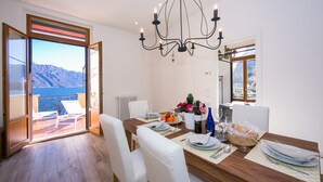 Ein Essbereich mit einem Holztisch, gedeckt für vier Personen, weißen Stühlen, einer Kristallleuchter und Blick auf das Meer durch eine Glastür.
