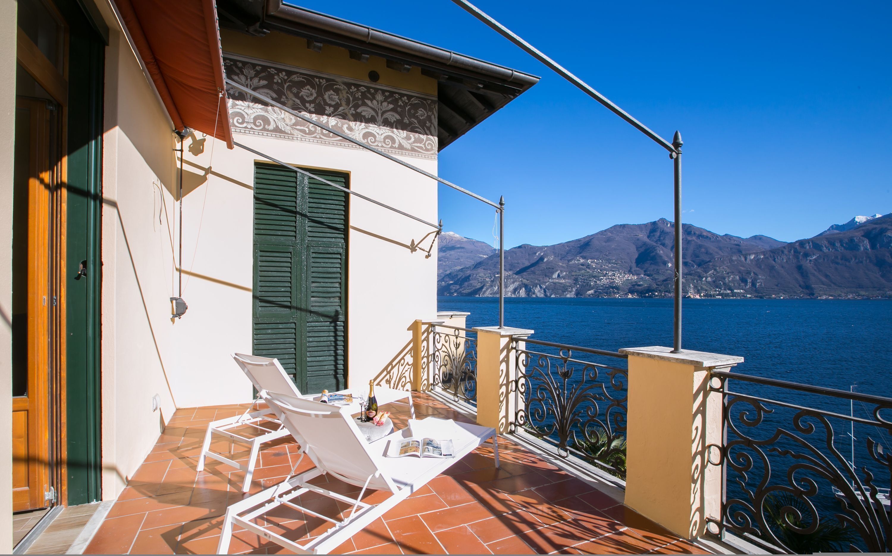 A balcony with white lounge chairs, a tiled floor, and a view of a lake with mountains in the background.