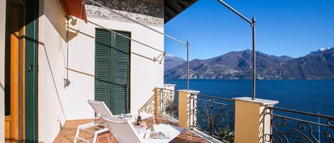 A balcony with white lounge chairs, a tiled floor, and a view of a lake with mountains in the background.