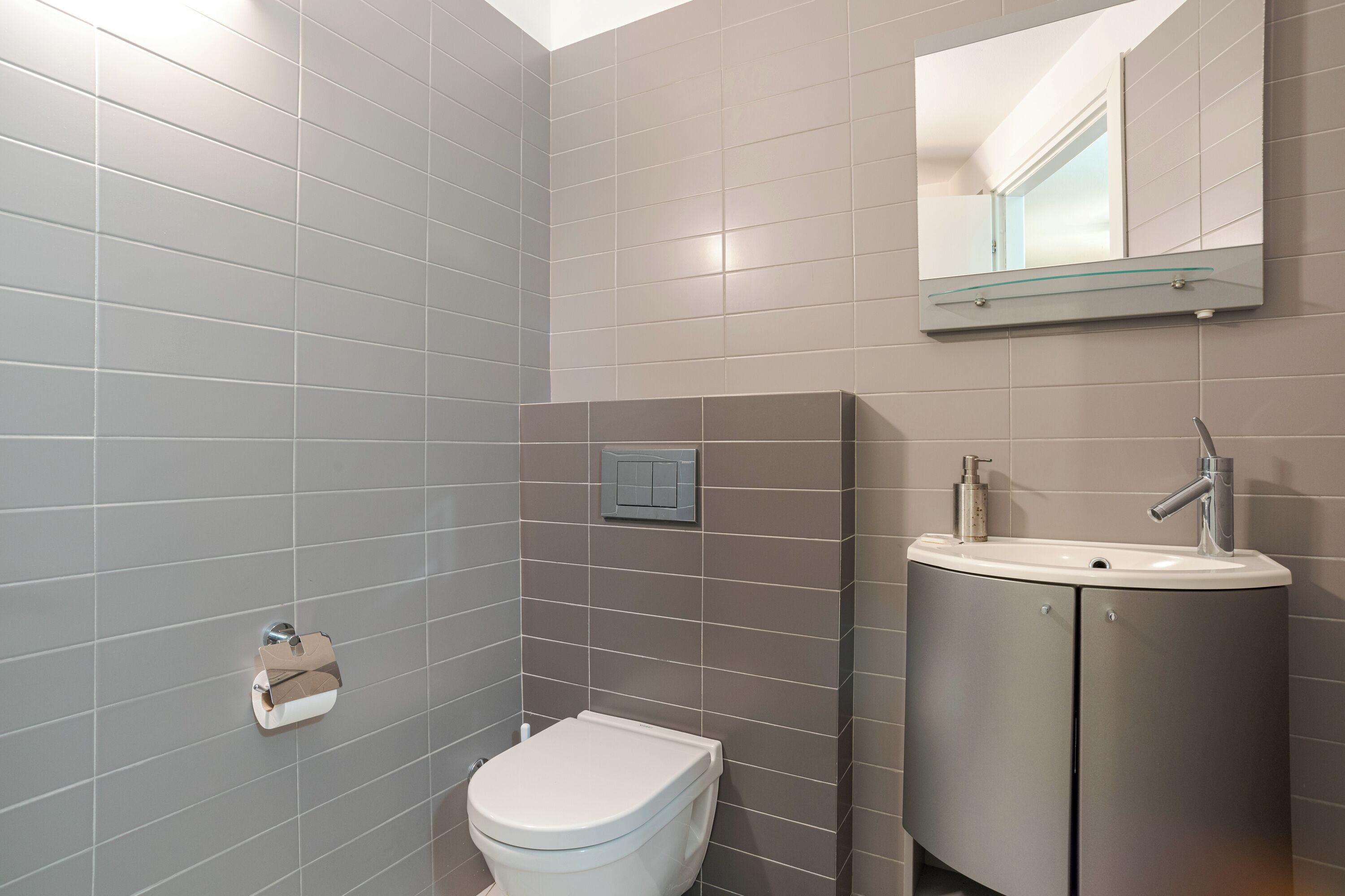 Ein modernes Badezimmer mit grauen Fliesen, einer Toilette, einem Waschbecken inklusive Spiegel und einem Seifenspender.