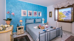 Quarto com parede azul, cama com estrutura de metal, mesa de cabeceira com abajur, cômoda e janela com vista para as montanhas.