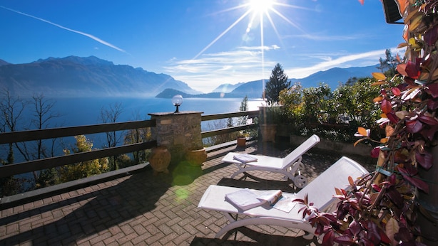 Um terraço com duas poltronas brancas, uma mesa de pedra e vista para montanhas e um lago.