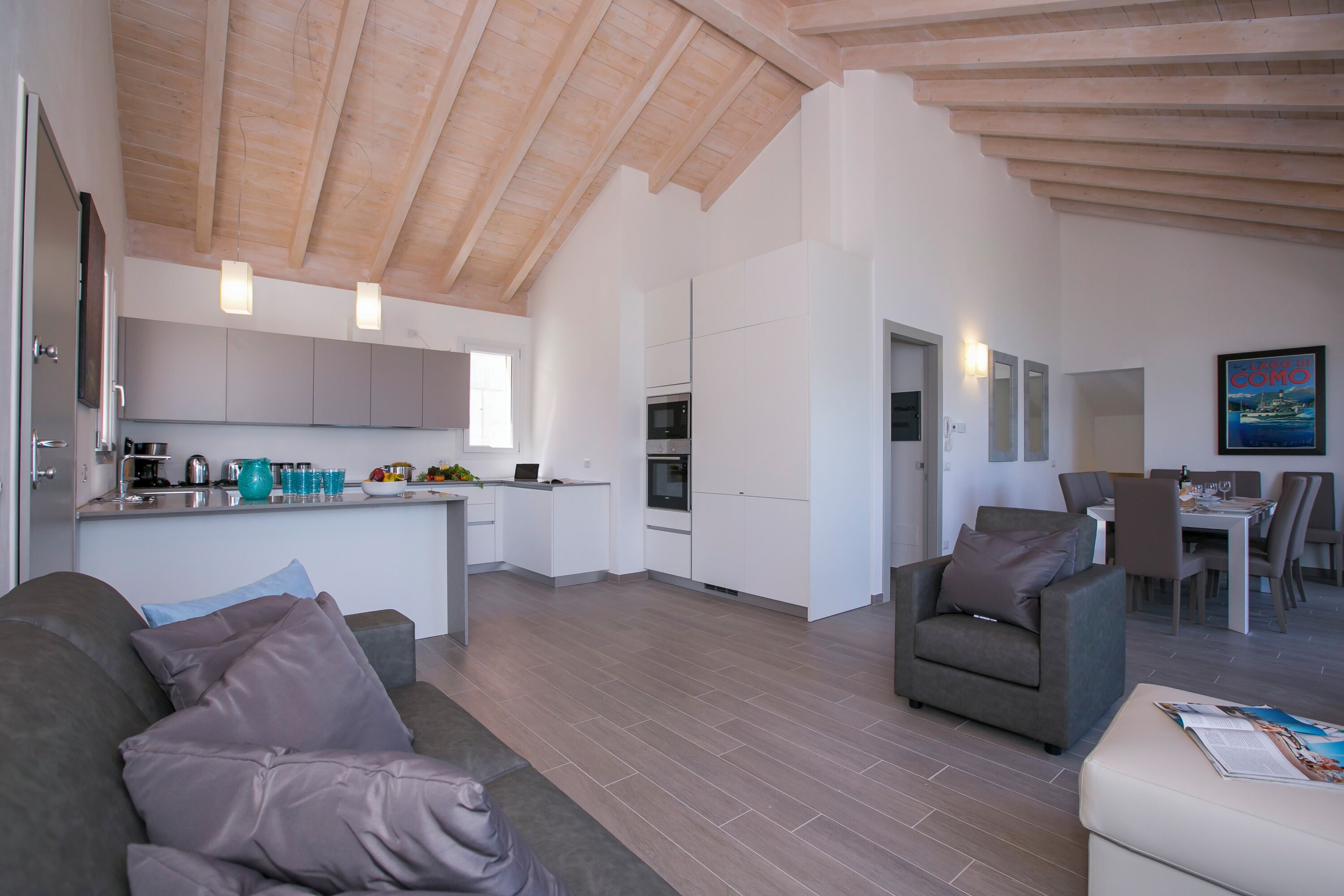 A modern kitchen with white cabinets, a dining area with a table and chairs, and a living room with a sofa and cushions.