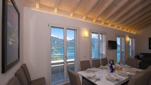 A dining area with a table set for four, large windows offering a view of mountains and a body of water, and a ceiling with exposed wooden beams. - Paradiso di Menaggio Penthouse (Menaggio)