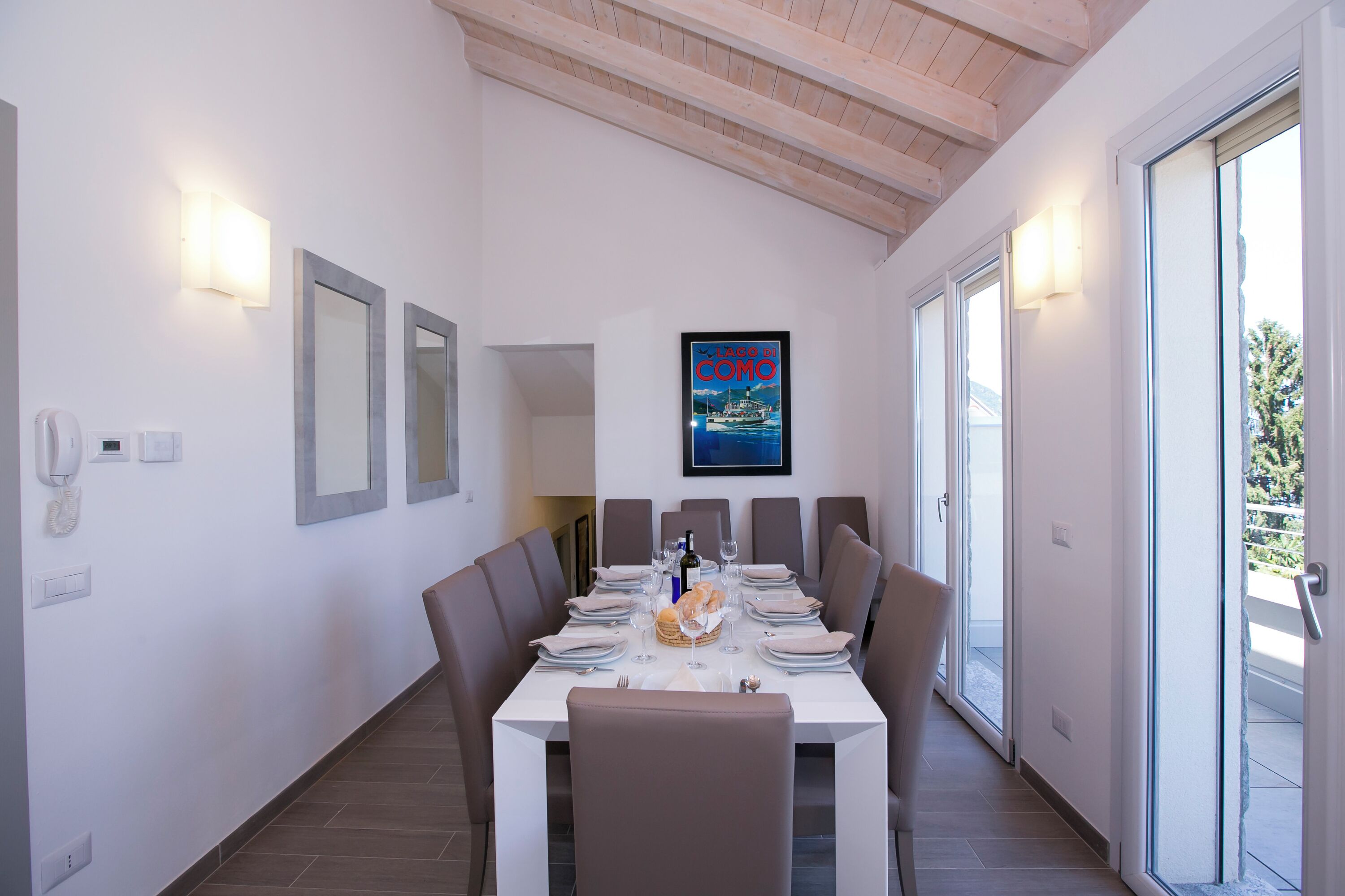 A dining area with a white table set for six, surrounded by chairs, with a framed poster on the wall and a phone on the wall.
