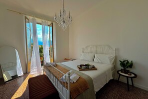 A bedroom with a bed, a bedside table with a plant, a chandelier, and a view of the outdoors. - Giardino Floreale di Menaggio (Menaggio)