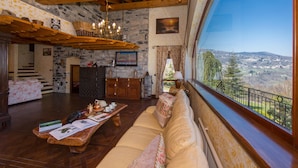 Ein gemütliches Interieur mit Steinwand, Holzbalken, einem großen Fenster mit schöner Aussicht, einer Couch und einem Holztisch mit Dekoration.