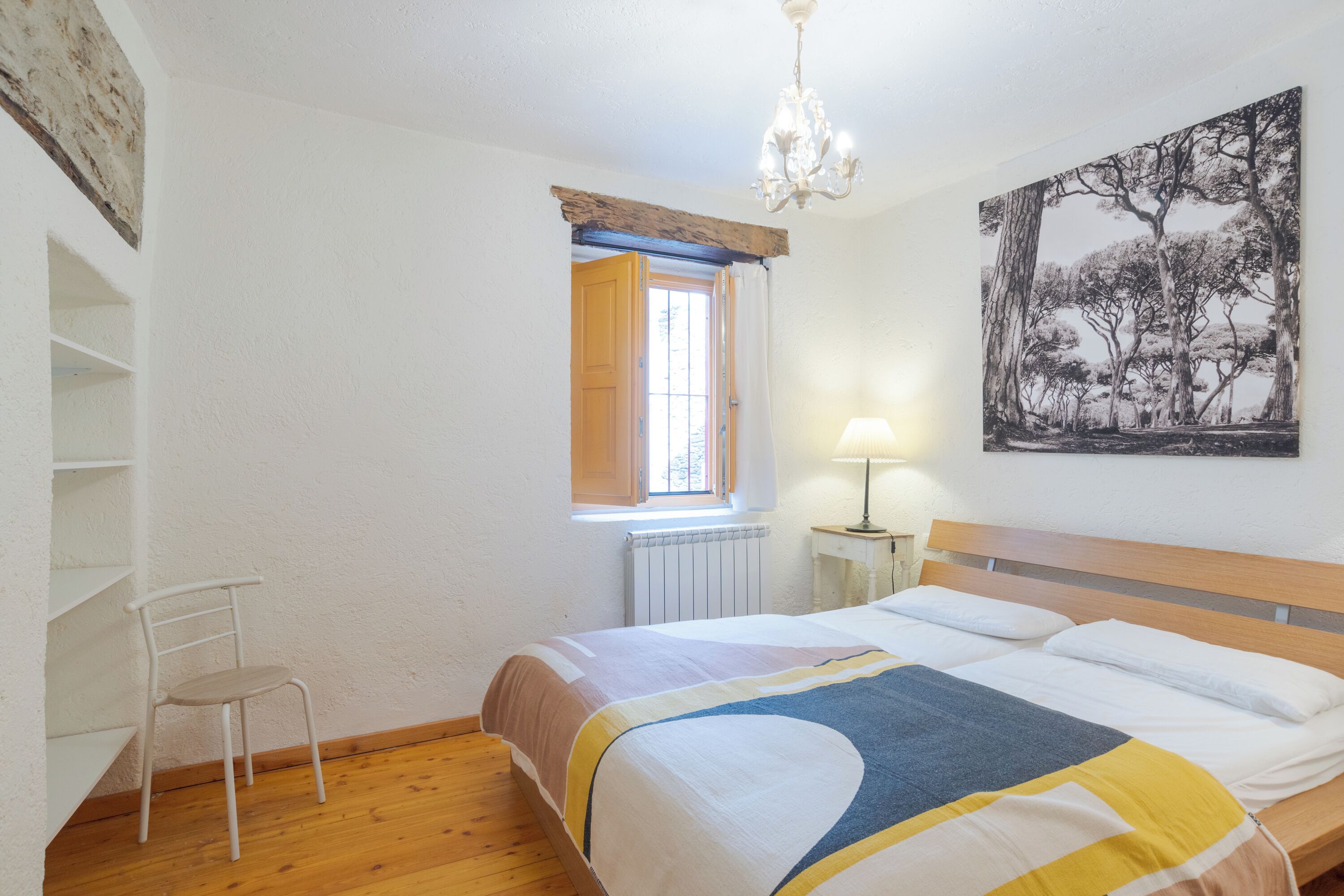 Ein Schlafzimmer mit einem hölzernen Bett, einem weißen Stuhl, einem Fenster mit Vorhängen, einem Heizkörper und einer Chandeliebre.