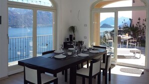 A dining area with a table set for four, large arched windows, and a view of the sea and mountains. - Villa Panoramica in Menaggio (Plesio)