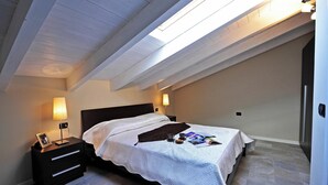 Une chambre avec une lucarne, un lit recouvert d’une courtepointe blanche, une table de chevet avec une lampe et un magazine posé sur le lit.