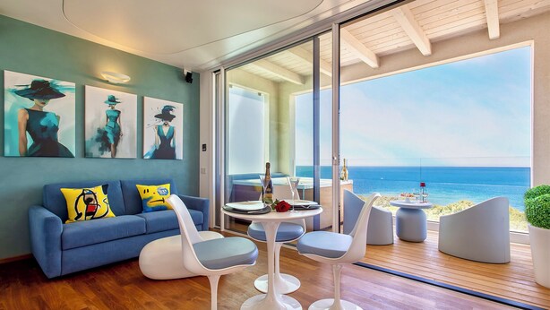 Una sala de estar moderna con un sofá azul, sillas blancas y una mesa lista para comer. Hay cuadros en la pared y vistas al mar a través de amplias ventanas.