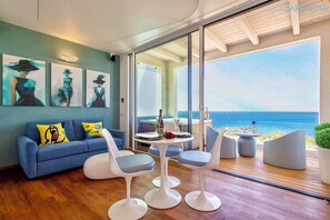 A modern living room with a blue sofa, white chairs, and a table set for dining. There are paintings on the wall and a view of the sea through large windows. - ClickSardinia Amare Penthouse with Hot Tub on Sea Front Terrace  (Alghero)