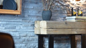 Un'atmosfera rustica con un muro in pietra, uno scaffale di legno con piatti, un vaso con rami e un tavolo con libri.