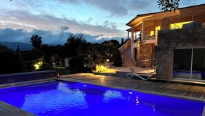 Ein Schwimmbecken mit Holzterrasse und Liegestühlen, umgeben von einer Steinmauer und einem modernen Haus mit Balkon.
