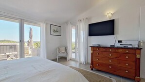 Ein Schlafzimmer mit einem großen Bett, einer hölzernen Kommode, einem Flachbildfernseher und einer Glastür, die auf einen Balkon mit Blick auf die Hügel führt.