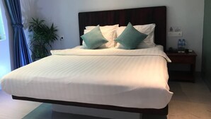 Un lit bien fait, avec du linge de lit blanc et deux coussins turquoise, une tête de lit en bois foncé, une table de chevet avec une lampe et une plante en pot.