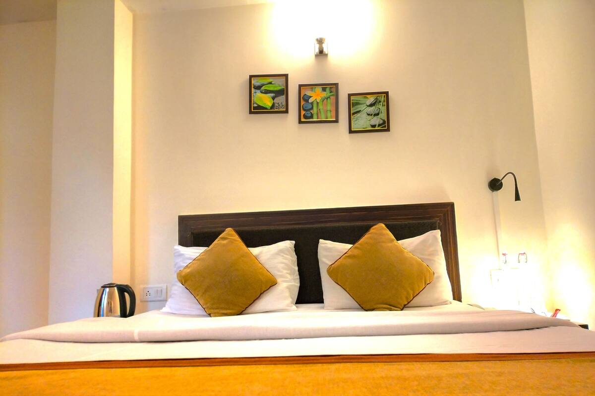 A hotel room with a large bed, two pillows, a bedside table with a kettle, and three framed pictures on the wall.