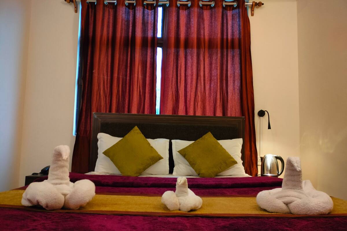 A hotel room with a bed, two pillows, a nightstand, a kettle, and a towel on the bed.