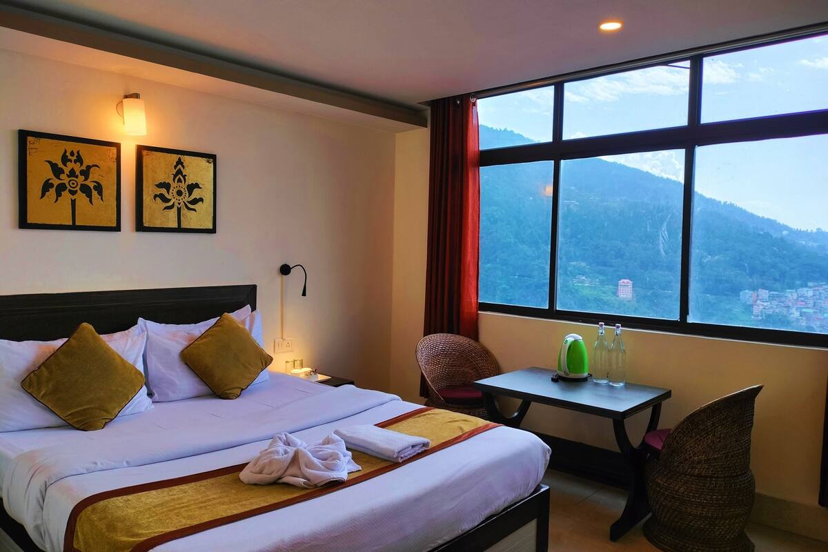 A hotel room with a bed, a nightstand, a table with a kettle and bottles, a chair, and a large window with a mountain view.
