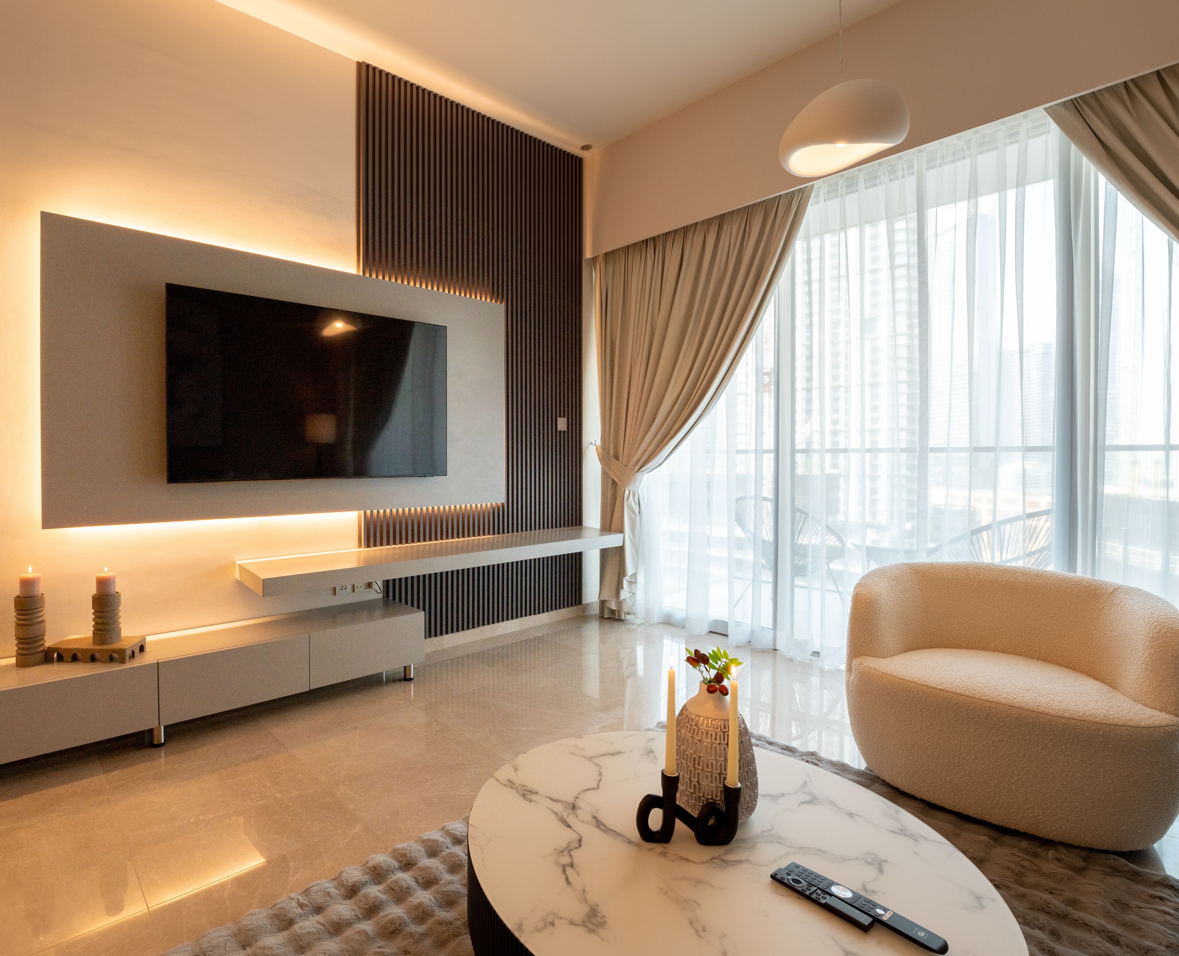 Een moderne woonkamer met een flatscreen televisie aan de muur, een comfortabele bank, een marmeren salontafel en een groot raam met dunne gordijnen.
