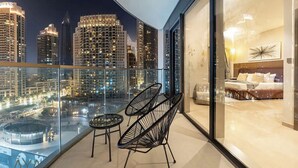 Een balkon met uitzicht op de stad, twee zwarte draadframe stoelen, en een blik op een slaapkamer met een bed en nachtkastje.