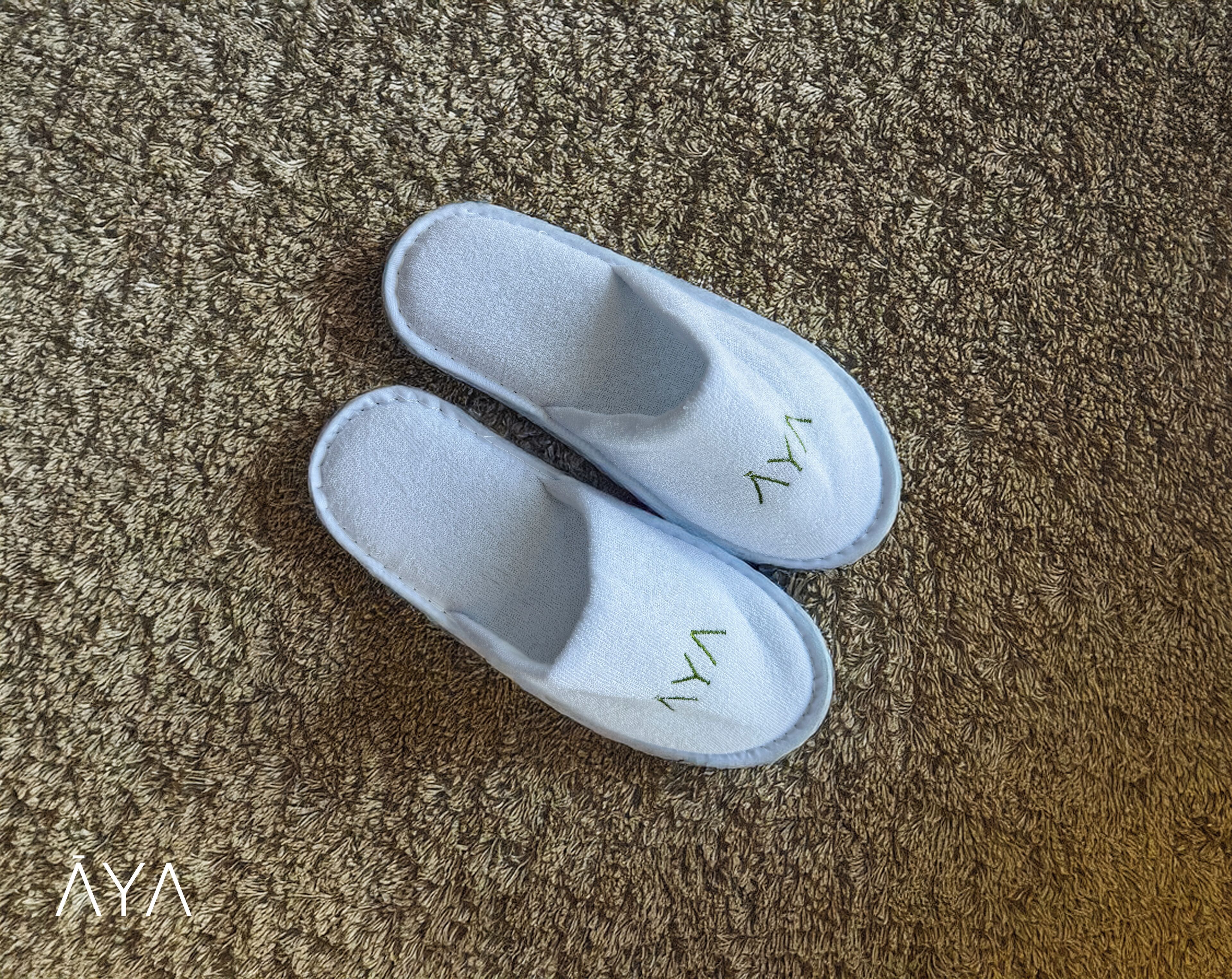 A pair of white slippers with embroidered letters on the sole, placed on a textured carpet.