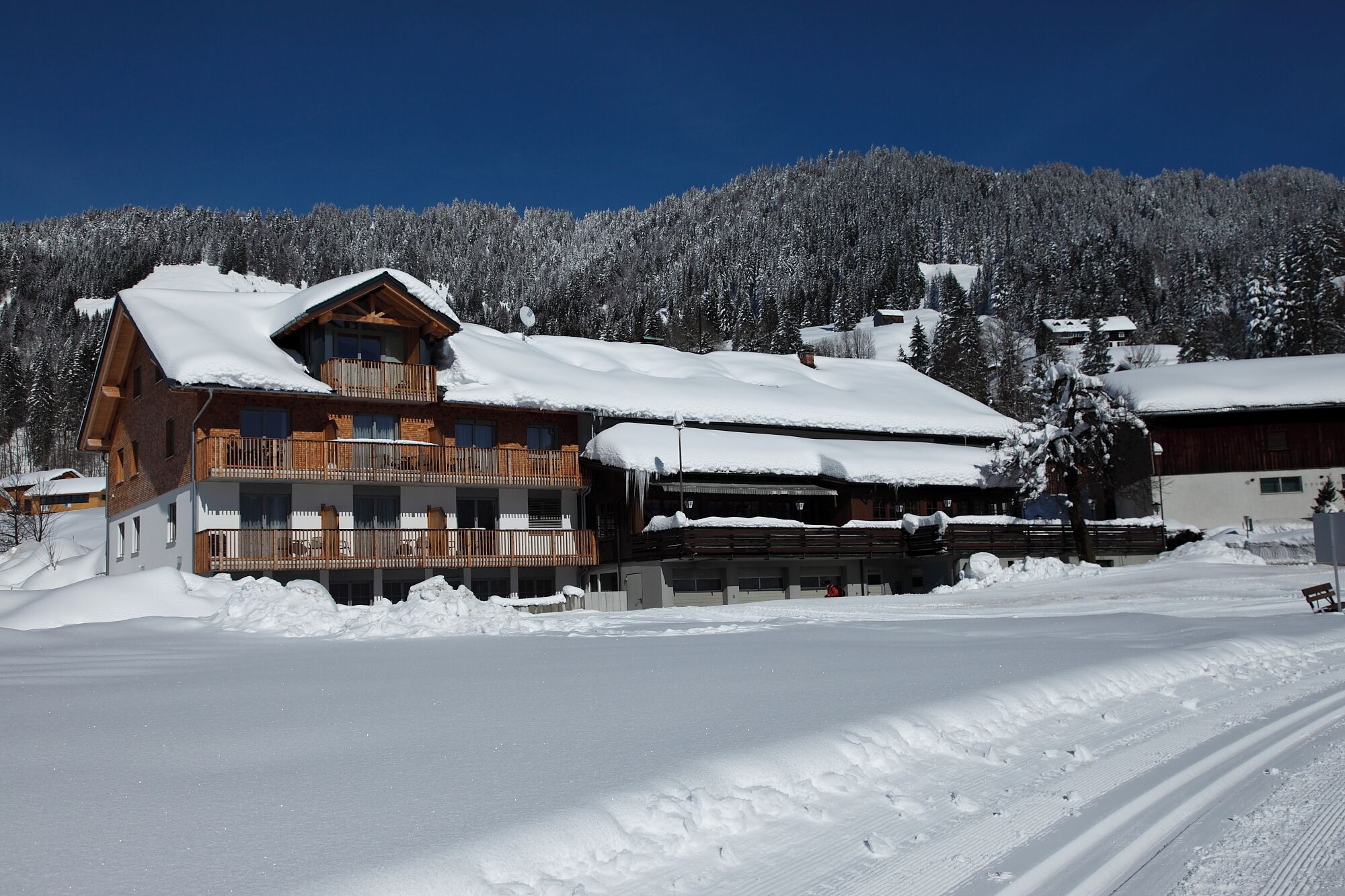 A wooden chalet with a balcony, surrounded by snow-covered trees and a clear blue sky.