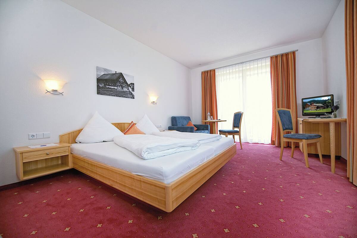Une chambre d’hôtel avec un lit, un bureau avec un ordinateur, deux chaises, une petite table et une fenêtre avec des rideaux.