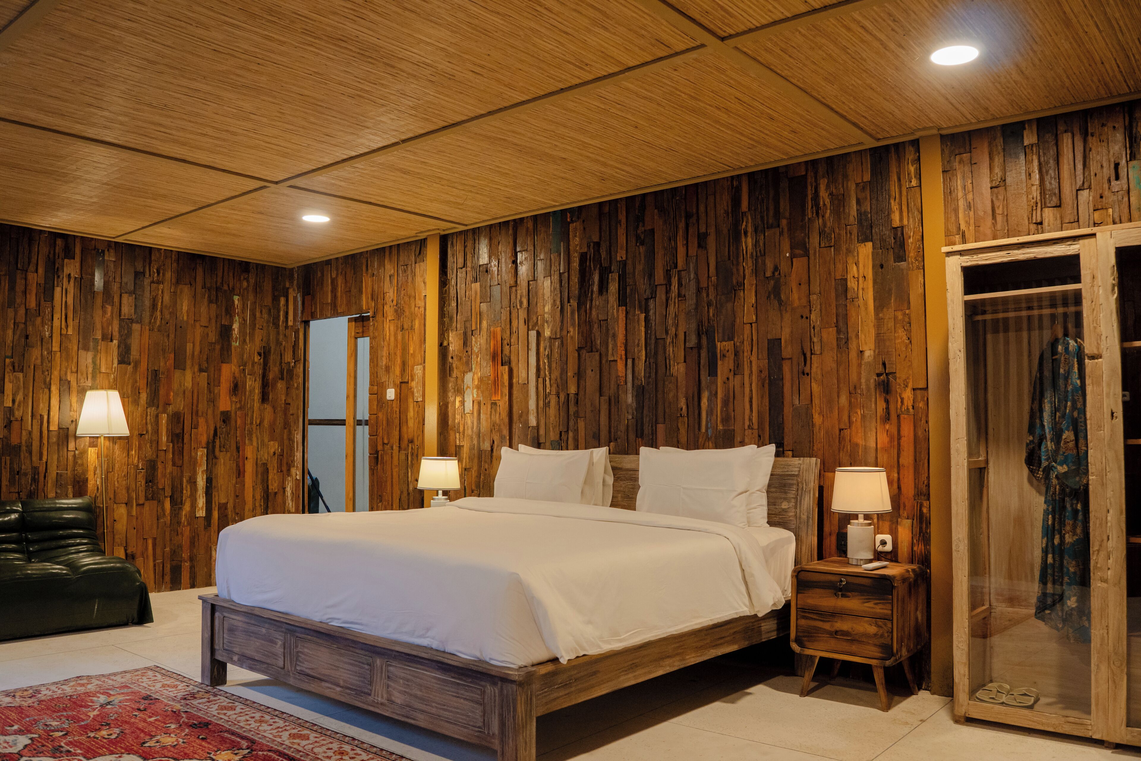 Un dormitorio con un panel de madera, una cama, una mesita de noche, una lámpara, un espejo y una puerta corrediza.