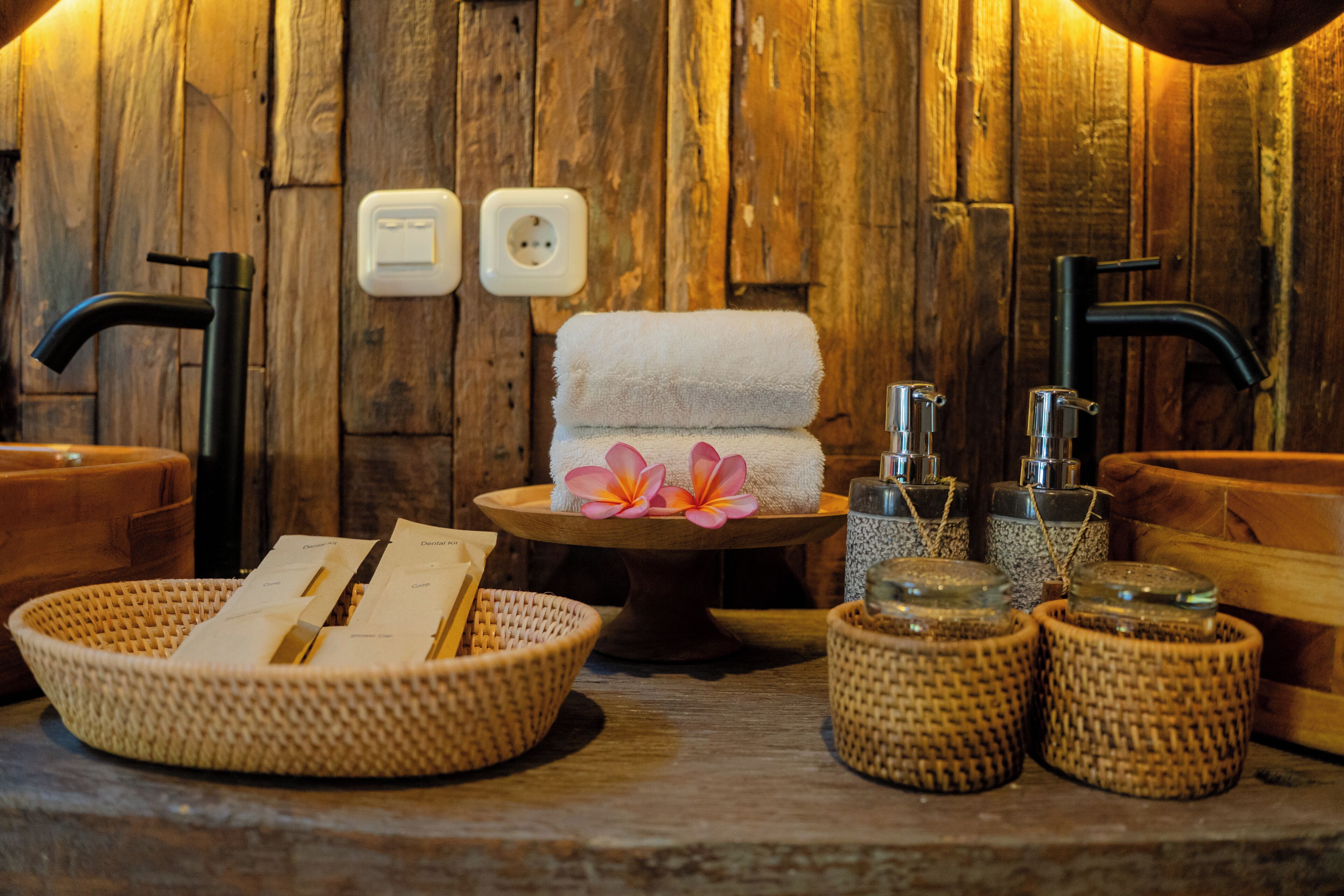 A wooden spa area with two black faucets, a white towel, a flower, and baskets of toiletries.