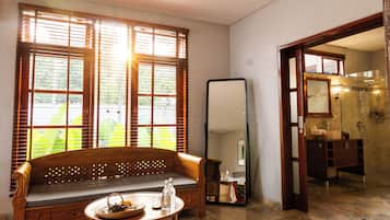 Una habitación con un banco de madera, una mesa redonda con tetera, un espejo grande y un baño visible a través de una puerta abierta.