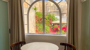 A room with a round table and two chairs, a large arched window with a view of a garden and stone buildings. - Marina View - Front of Sea Cospicua (Cospicua)