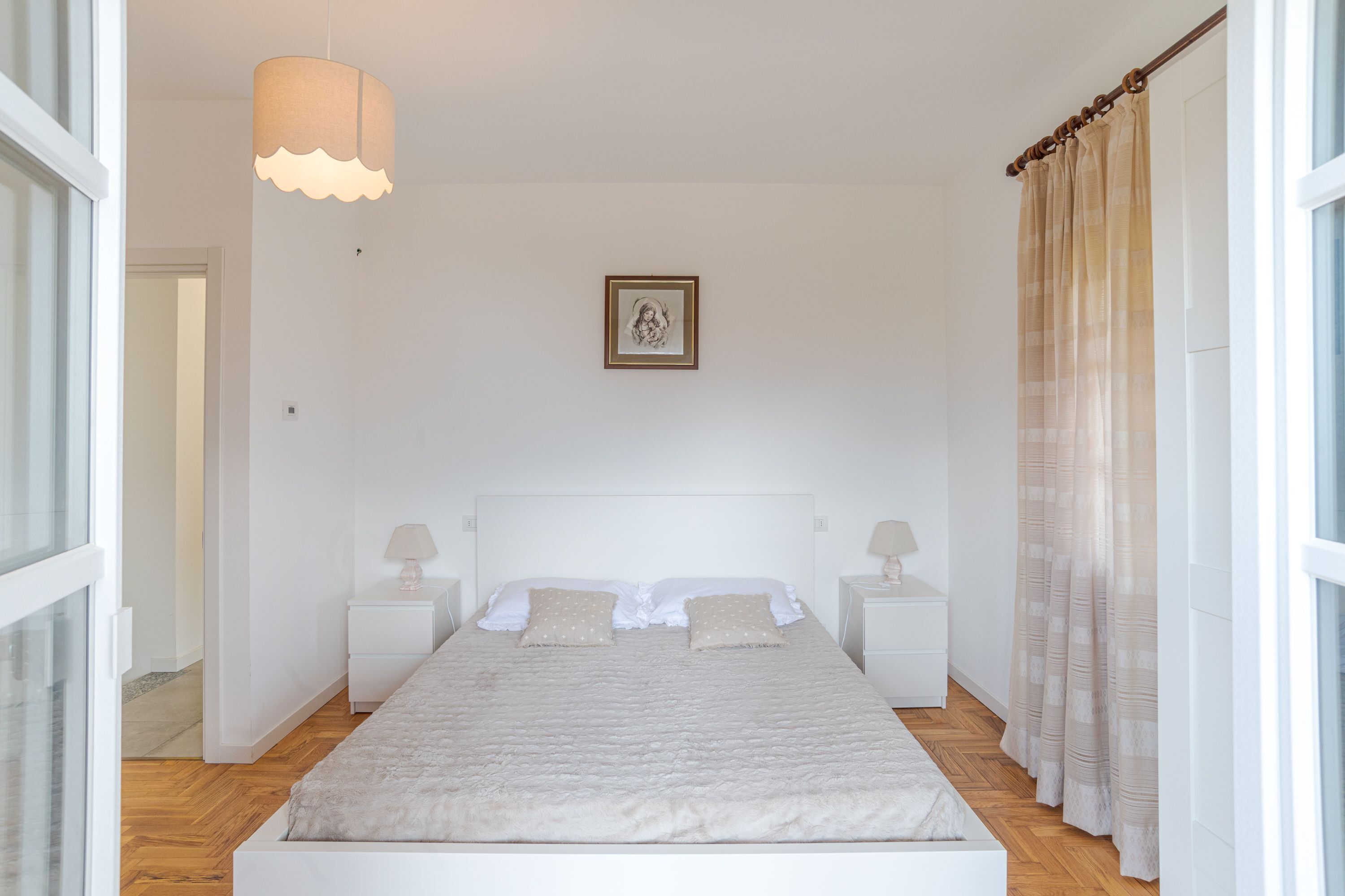 Ein Schlafzimmer mit einem großen Bett, zwei Nachttischen, einer Pendelleuchte, einem gerahmten Bild an der Wand und einem Fenster mit Vorhängen.