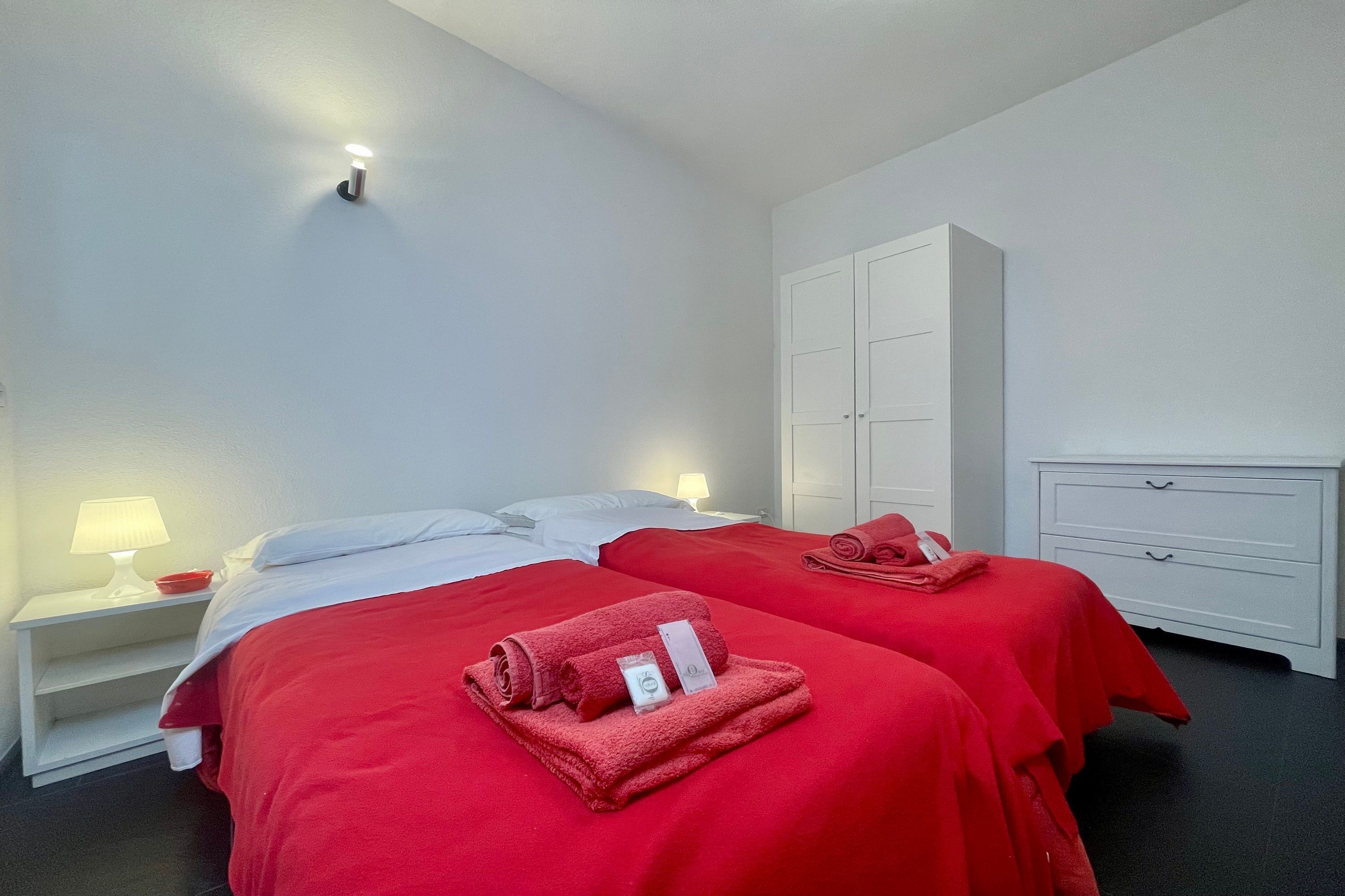 A hotel room with two beds, each with a red blanket and a rolled towel. There is a white wardrobe and a nightstand with a lamp.