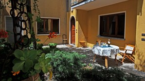 Une cour avec une table et des chaises, entourée de plantes et d’un bâtiment jaune doté de fenêtres et d’une porte.