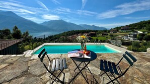 A poolside area with a view of mountains and a lake. There are two chairs and a table with a magazine and flowers. - Belmonte Terrazzo in Calozzo (Pianello del Lario)