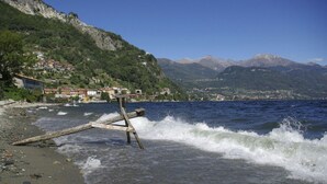 A wooden structure on a rocky shoreline with waves crashing against it. In the background, there are buildings on a hillside and mountains in the distance. - Belmonte Terrazzo in Calozzo (Pianello del Lario)