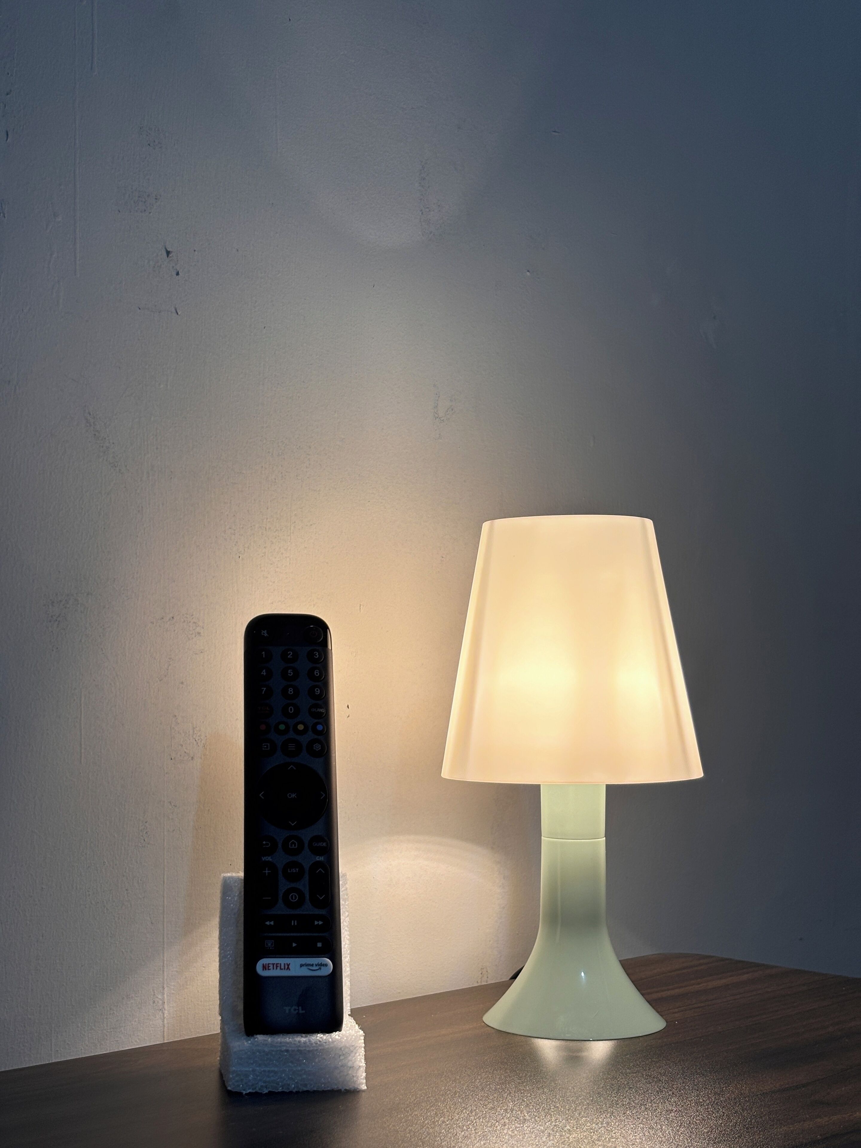A remote control on a foam holder and a table lamp on a wooden surface.