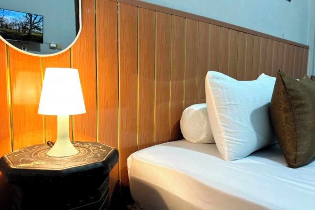 A neatly made bed with white and brown pillows, a bedside lamp, and a mirror on a wooden panel wall.