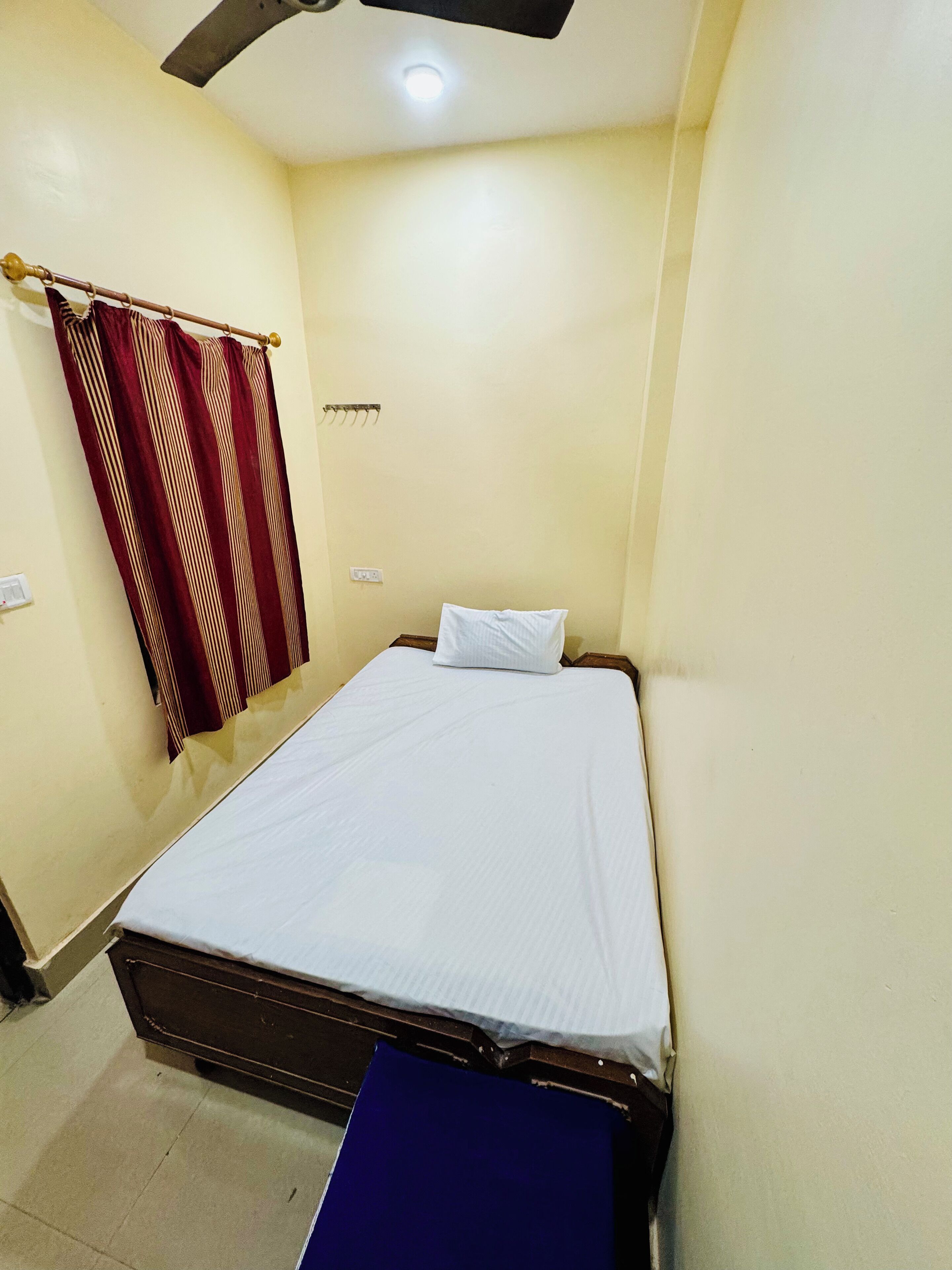 A single bed with white bedding in a room with cream walls, a ceiling fan, and a curtain with a striped pattern.