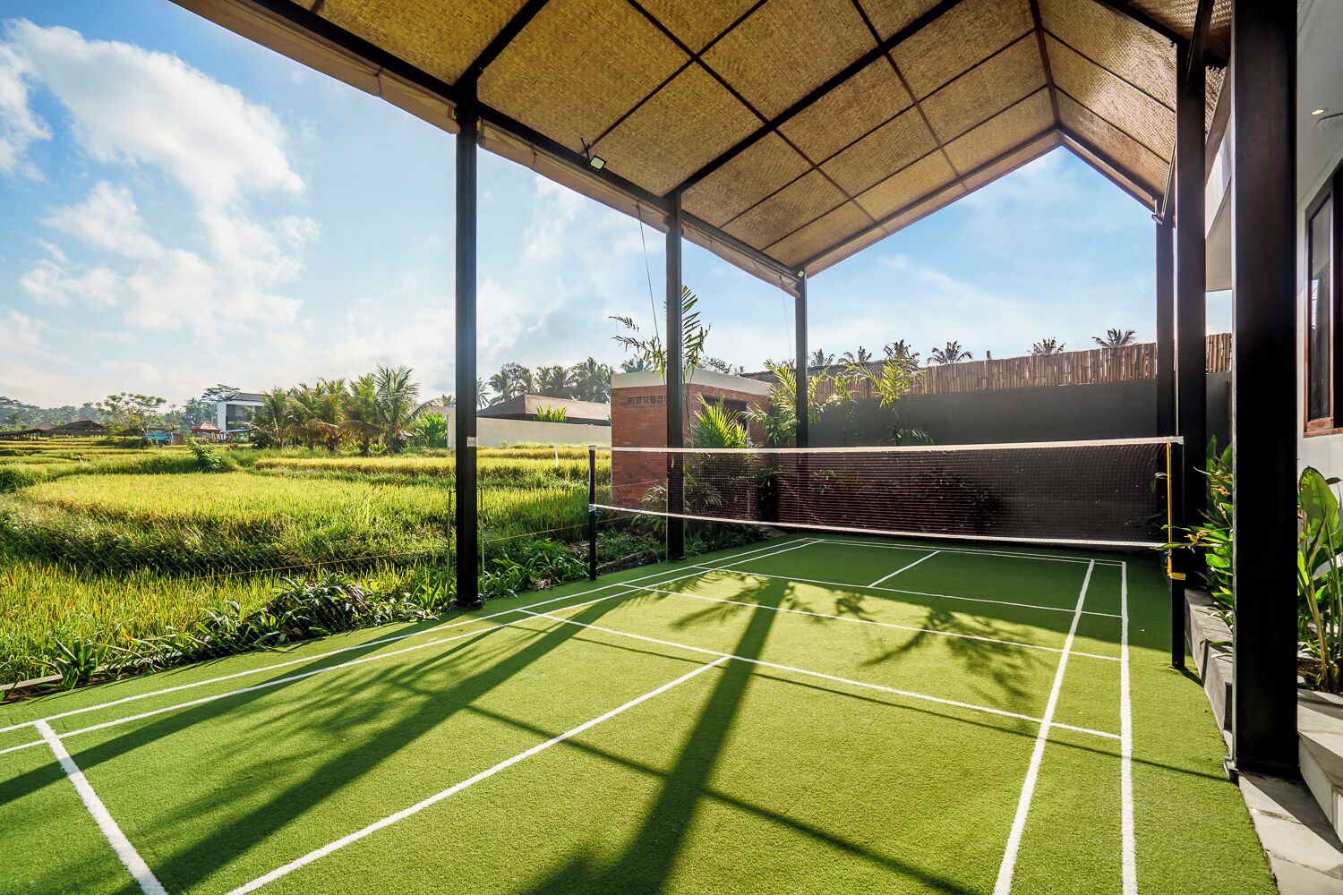 A tennis court with a net, surrounded by a covered area with a view of a rice field and a building in the background.