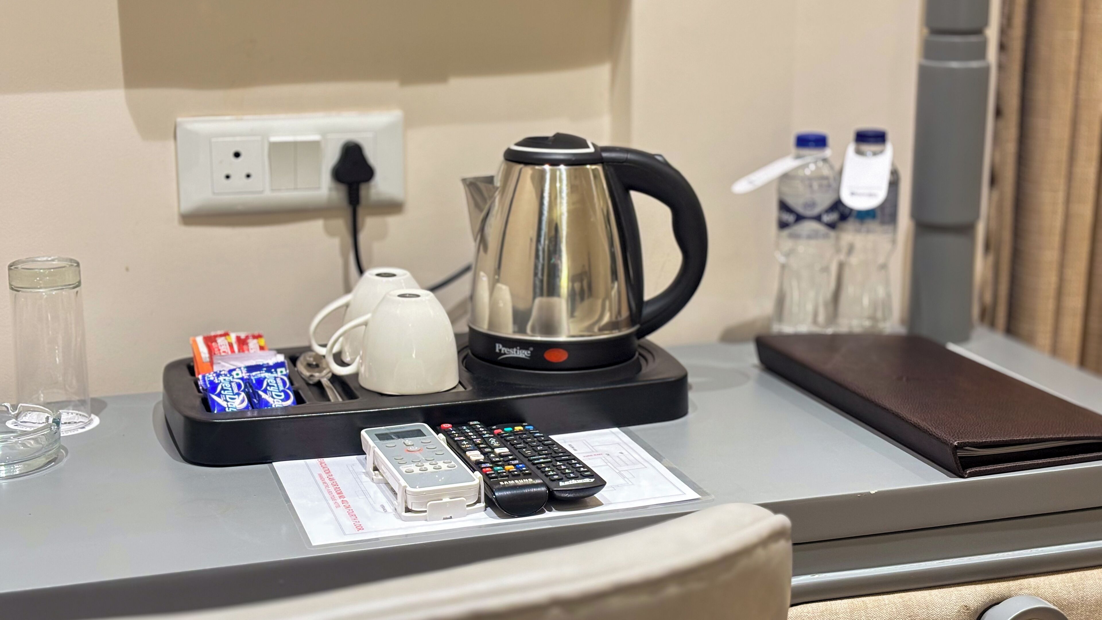 A hotel room desk with a kettle, two cups, a glass, a remote control, and a mobile phone.
